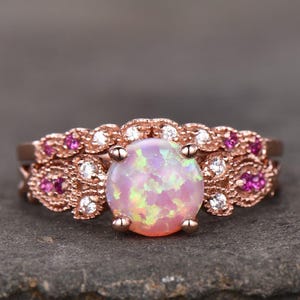 May include: Rose gold ring featuring a large, round opal gemstone with pink and green hues. The band is adorned with small, round pink gemstones and clear, round gemstones. The ring is set against a dark gray background.