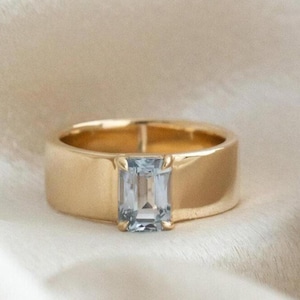 Peut inclure: Une bague en or avec une pierre précieuse rectangulaire bleu clair. La bague a un anneau épais et une finition polie. La pierre est sertie au centre, créant un design simple mais élégant. La bague est présentée sur une surface blanche texturée.