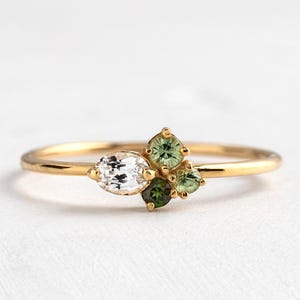 Può includere: Un delicato anello d'oro con un gruppo di pietre preziose. L'anello presenta una gemma trasparente taglio marquise e un gruppo di gemme verdi. L'anello è montato su una sottile fascia d'oro.