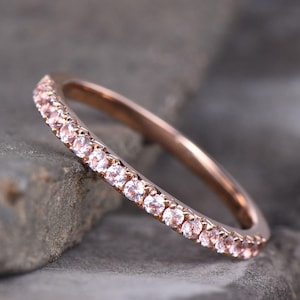 May include: Rose gold ring with a band of small, light pink gemstones. The ring is set against a gray, textured background, showcasing the delicate details of the jewelry. The gemstones are evenly spaced around the band.