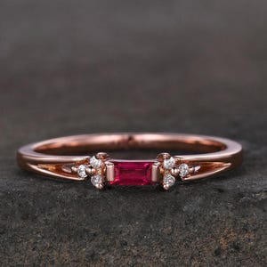 May include: Rose gold ring with a central rectangular, vivid red gemstone, and small clusters of white gemstones on either side. The band has a delicate, openwork design. A piece of jewellery.