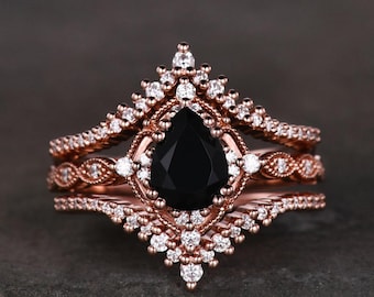 May include: A rose gold ring set featuring a pear-shaped black gemstone at the center. The engagement ring is surrounded by a halo of small white gemstones, with additional gemstone accents on the bands.