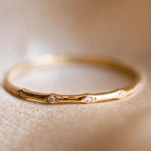 Pode incluir: Um delicado anel de ouro com pequenas pedras preciosas brilhantes, espaçadas uniformemente ao redor da banda. O anel tem um acabamento liso e polido e é colocado sobre um fundo macio e neutro. Esta elegante joia é perfeita para o uso diário.