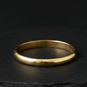 May include: A gold ring with a smooth, rounded band. The ring is displayed on a dark surface, accentuating its polished finish. The ring is a simple, elegant piece of jewellery.