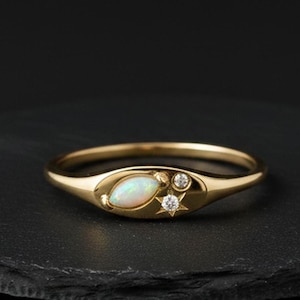 May include: A gold ring with an oval opal center stone, flanked by a small diamond and a star-shaped accent. The ring is set on a dark, textured surface, showcasing its delicate design and craftsmanship. The ring's band is thin and polished.