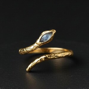 May include: A gold-toned open ring featuring a textured, branch-like band and a single marquise-cut, iridescent blue stone set at one end. The metal surface has a rough, organic finish, and the ring is shaped in a bypass style.