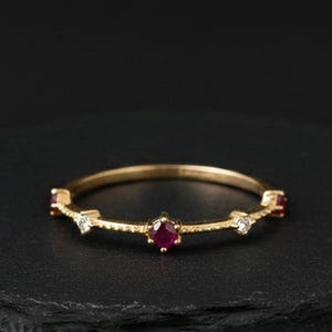 Puede incluir: Un delicado anillo de oro con un rubí central, flanqueado por piedras blancas más pequeñas y rubíes. El anillo está colocado sobre una superficie negra, mostrando su intrincado diseño y colores vibrantes. Una joya elegante para cualquier ocasión.