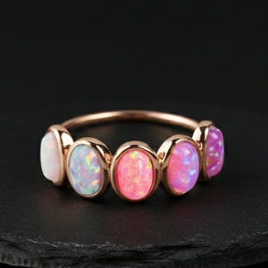 May include: A rose gold ring featuring five oval-shaped opal stones in shades of white, blue, pink, and purple. The ring is set on a black surface, showcasing the colorful gemstones.