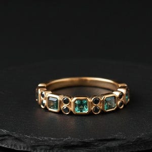 Op de afbeelding: Een gouden ring met afwisselende vierkante turkooizen en ronde zwarte edelstenen. De ring wordt gepresenteerd op een donker, getextureerd oppervlak, waarbij het herhalende patroon wordt benadrukt.