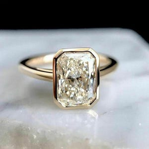May include: A gold ring with a large, rectangular, light-coloured gemstone. The gemstone is set within a gold bezel, and the band is a simple, polished gold. The ring is displayed on a white surface.
