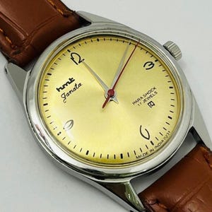 May include: A vintage HMT Janata wristwatch with a gold-coloured face, silver case, and brown leather strap. The watch face features black numerals, the words "hmt Janata", and "PARA SHOCK 17 JEWELS". The watch has a red second hand.