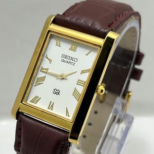 May include: A rectangular Seiko Quartz wristwatch featuring a gold-tone case, a white dial with gold Roman numeral hour markers, and a dark burgundy leather strap with a textured, embossed pattern and a gold-colored side crown.