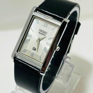 May include: A rectangular Seiko Quartz wristwatch featuring a silver-toned dial with Roman numeral hour markers, a textured center pattern, and a black leather strap. The watch case is polished silver-tone metal with a small crown on the side.