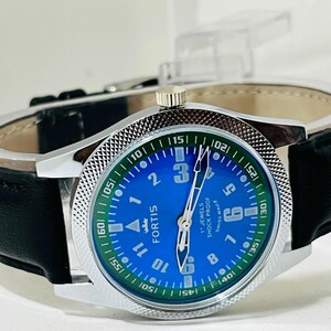 May include: A Fortis wristwatch with a bright blue dial, white Arabic numerals, and a green outer ring featuring white minute markers. The watch has a silver-tone metal case with a textured bezel, silver hands, and a black leather strap.