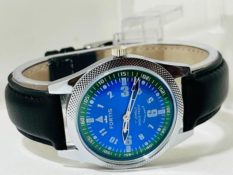 May include: A Fortis wristwatch with a bright blue dial, white Arabic numerals, and a green outer ring featuring white minute markers. The watch has a silver-tone metal case with a textured bezel, silver hands, and a black leather strap.