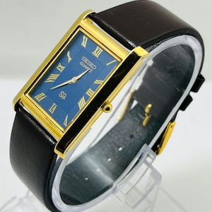 May include: A rectangular Seiko Quartz wristwatch featuring a gold-tone case, a deep blue dial with a textured wave pattern, and gold Roman numeral hour markers. The watch is fitted with a smooth black leather strap and a gold-tone buckle.