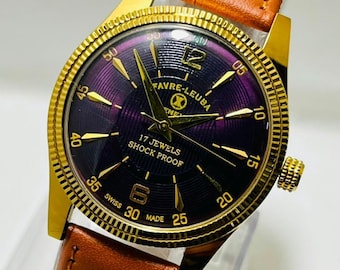 May include: A gold-tone wristwatch with a deep purple, textured dial featuring gold-tone hands and hour markers. The watch face displays the text FAVRE-LEUBA, 17 JEWELS, SHOCK PROOF, and SWISS MADE. It includes a brown leather strap with visible stitching.