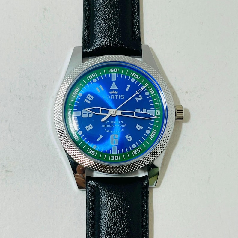 May include: A Fortis wristwatch with a blue dial, white Arabic numerals, and a green outer ring featuring white minute markers. The watch has a silver-toned textured bezel, silver hands, and a black leather strap. Text on the dial reads Fortis, 17 Jewels, Shockproof, and Swiss Made.