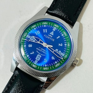 May include: A Fortis wristwatch with a blue dial, white Arabic numerals, and a green outer ring. The watch features silver hands, a textured silver bezel, and a black leather strap. The dial displays the text Fortis, 17 Jewels, Shock Proof, and Swiss Made.