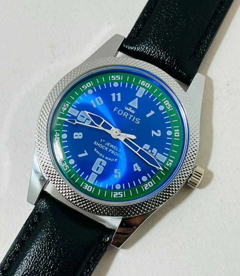 May include: A Fortis wristwatch with a blue dial, white Arabic numerals, and a green outer ring. The watch features silver hands, a textured silver bezel, and a black leather strap. The dial displays the text Fortis, 17 Jewels, Shock Proof, and Swiss Made.