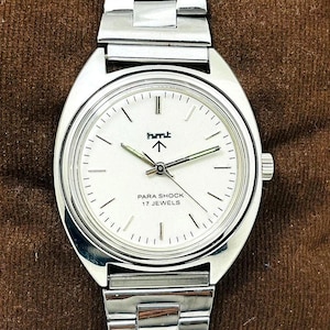 May include: A silver-toned wristwatch with a white face and a silver band. The watch face features the text "hmt" above an arrow, and "PARA SHOCK 17 JEWELS" below. The watch is displayed on a brown surface.