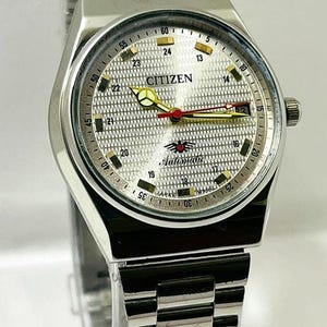 Puede incluir: Reloj automático Citizen plateado con esfera plateada texturizada. El reloj tiene una correa plateada y una pantalla de fecha. La esfera incluye el nombre de la marca y la palabra "Automatic".