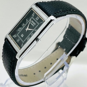 Puede incluir: Un reloj Seiko Quartz rectangular con esfera negra y caja plateada. El reloj presenta números romanos y una correa de cuero negro. El reloj se exhibe sobre un soporte transparente.