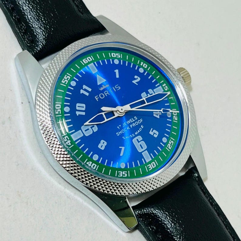 May include: A Fortis wristwatch with a blue dial, white Arabic numerals, and a green outer ring featuring white minute markers. The watch has a silver-toned textured bezel, silver hands, and a black leather strap. The dial displays 17 JEWELS SHOCK PROOF.