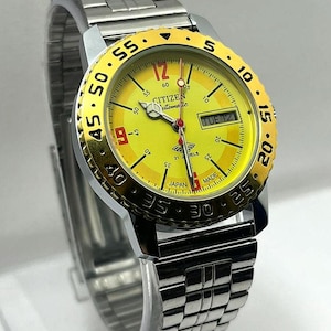 May include: A Citizen automatic wristwatch with a yellow face and a gold-coloured bezel. The watch has a silver stainless steel strap and shows the day and date. The dial includes the text "Citizen Automatic" and "Made in Japan".