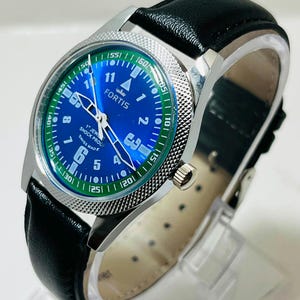 May include: A Fortis wristwatch with a blue dial, white Arabic numerals, and a green outer ring. The watch features silver hands, a textured silver bezel, and a black leather strap. The dial text reads Fortis, 17 Jewels, Shock Proof, Swiss Made.