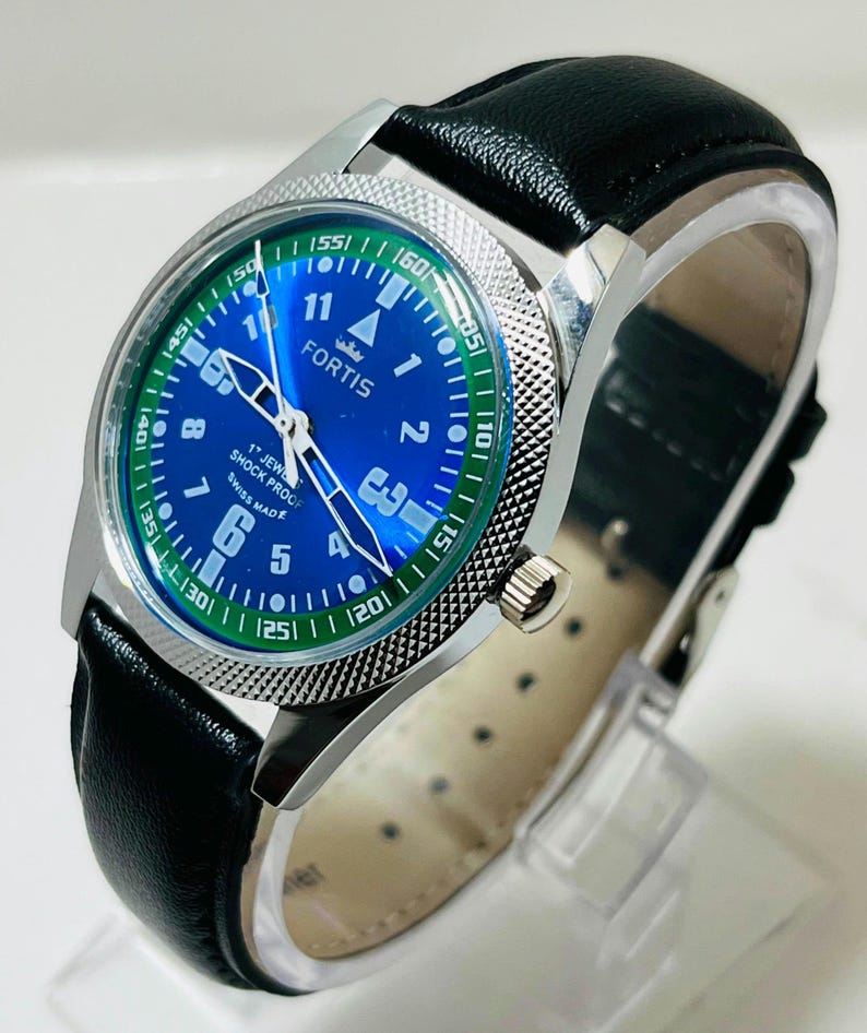 May include: A Fortis wristwatch with a blue dial, white Arabic numerals, and a green outer ring. The watch features silver hands, a textured silver bezel, and a black leather strap. The dial text reads Fortis, 17 Jewels, Shock Proof, Swiss Made.