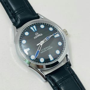 Peut inclure: Une montre Roamer argentée avec un cadran noir et un bracelet en cuir noir. Le cadran de la montre présente des accents blancs et bleus, avec le texte "17 Jewels Shock Proof". La montre a un design classique et sportif.
