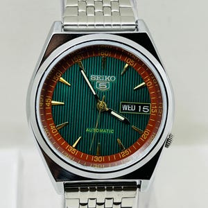 May include: A silver-toned Seiko 5 automatic wristwatch. The watch features a green dial with gold-toned hands and markers, a day-date display, and a stainless steel band. The bezel has a red and black outer ring.