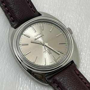 May include: A silver wristwatch with a light silver dial and a dark brown leather strap. The watch face displays the brand name "HMT Kohinoor" and the text "PARA SHOCK 17 JEWELS". The watch has a round face and a crown on the side.