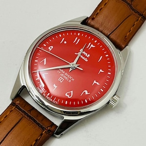 May include: A vintage wristwatch with a red face and Arabic numerals. The watch has a silver-coloured case and a brown leather strap. The watch face displays the text "ARA SHOCK 17 JEWELS" and "MADE IN INDIA 0231".