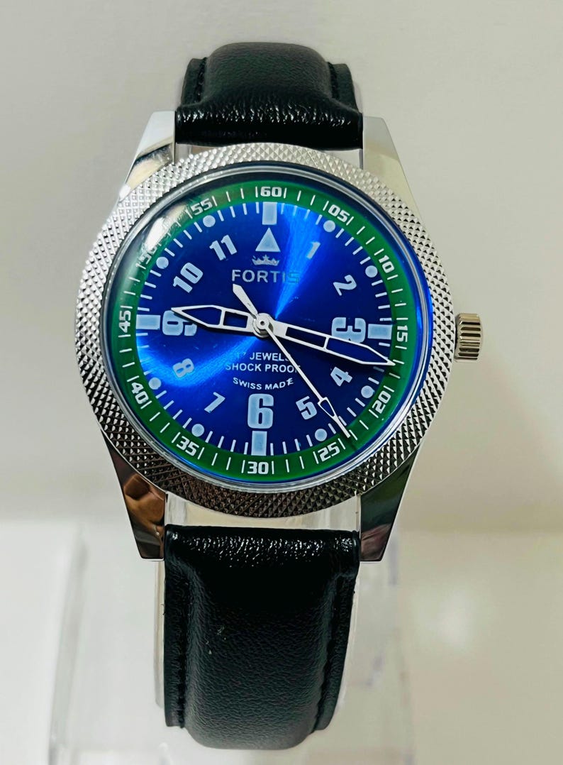 May include: A Fortis wristwatch with a blue sunray dial, white Arabic numerals, and luminous hands. The watch features a silver-toned textured bezel, a black leather strap, and text on the dial reading FORTIS, JEWELED, SHOCK PROOF, and SWISS MADE.