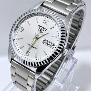 May include: A silver-tone Seiko 5 automatic wristwatch featuring a white dial with silver indices, Arabic numerals at 6, 9, and 12 o'clock, a day-date window at 3 o'clock, luminous hands, a fluted bezel, and a brushed stainless steel link bracelet.