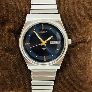 Puede incluir: Un reloj Citizen plateado con esfera azul oscuro y agujas e indicadores dorados. El reloj muestra "CITIZEN" en la parte superior y "FRI 17" en una pequeña ventana. La correa es plateada, sobre un fondo marrón.