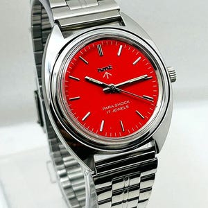 May include: A stainless steel wristwatch with a vibrant red face. The watch features a silver band and a round dial with white hour markers and hands. The text "PARA SHOCK 17 JEWELS" is visible on the watch face.