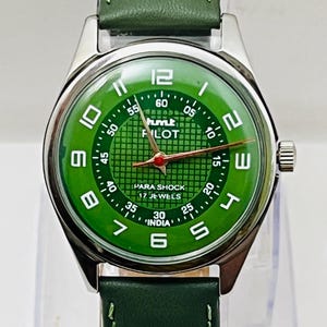 Może przedstawiać: Zegarek na rękę z zieloną tarczą, srebrną kopertą i zielonym skórzanym paskiem. Tarcza posiada białe cyfry, czerwoną wskazówkę sekundową oraz napis "hmt PILOT PARA SHOCK 17 JEWELS INDIA".