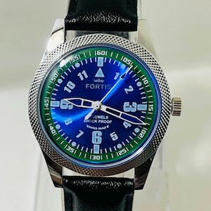 May include: A Fortis wristwatch with a blue sunray dial, white Arabic numerals, and a green outer chapter ring. The watch features a silver-tone textured bezel, luminous hands, and a black leather strap. Text on the dial reads FORTIS, JEWELS, SHOCK PROOF, and SWISS MADE.
