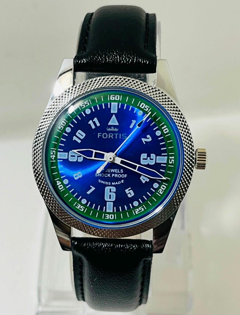 May include: A Fortis wristwatch with a blue sunray dial, white Arabic numerals, and a green outer chapter ring. The watch features a silver-tone textured bezel, luminous hands, and a black leather strap. Text on the dial reads FORTIS, JEWELS, SHOCK PROOF, and SWISS MADE.