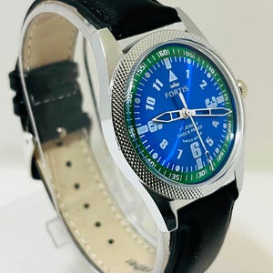 May include: A Fortis wristwatch with a blue dial, white Arabic numerals, and a green outer ring featuring white minute markers. The watch has a silver-tone metal case with a textured bezel and a black leather strap with a light-colored lining.