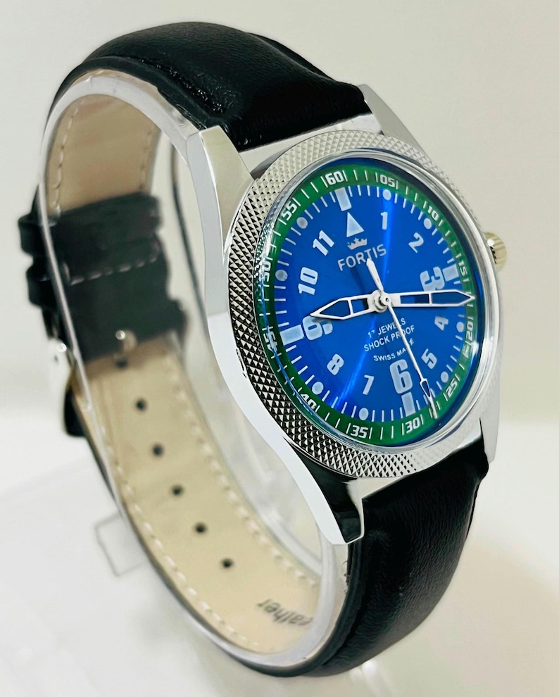 May include: A Fortis wristwatch with a blue dial, white Arabic numerals, and a green outer ring featuring white minute markers. The watch has a silver-tone metal case with a textured bezel and a black leather strap with a light-colored lining.