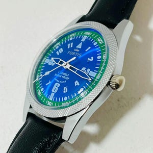 May include: A Fortis wristwatch with a blue dial, white Arabic numerals, and a green outer ring. The watch features a silver-toned textured bezel, silver hands, and a black leather strap. Text on the dial reads FORTIS, 17 JEWELS, SHOCK PROOF, SWISS MADE.