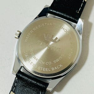 May include: A stainless steel watch case back with a black leather strap. The circular metal back features the engraved text "WATER RESISTANT," "SHOCK RESISTANT," "FORTIS," "FORTIS WATCH CO. SWISS," and "STAINLESS STEEL BACK" with a crown logo.