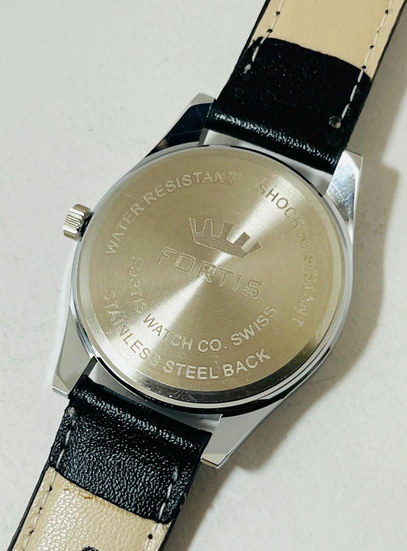 May include: A stainless steel watch case back with a black leather strap. The circular metal back features the engraved text "WATER RESISTANT," "SHOCK RESISTANT," "FORTIS," "FORTIS WATCH CO. SWISS," and "STAINLESS STEEL BACK" with a crown logo.