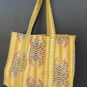 May include: A large tote bag with a yellow and orange striped pattern, featuring tiger illustrations. The bag has long straps and is made of a quilted fabric. The bag is designed for carrying items.