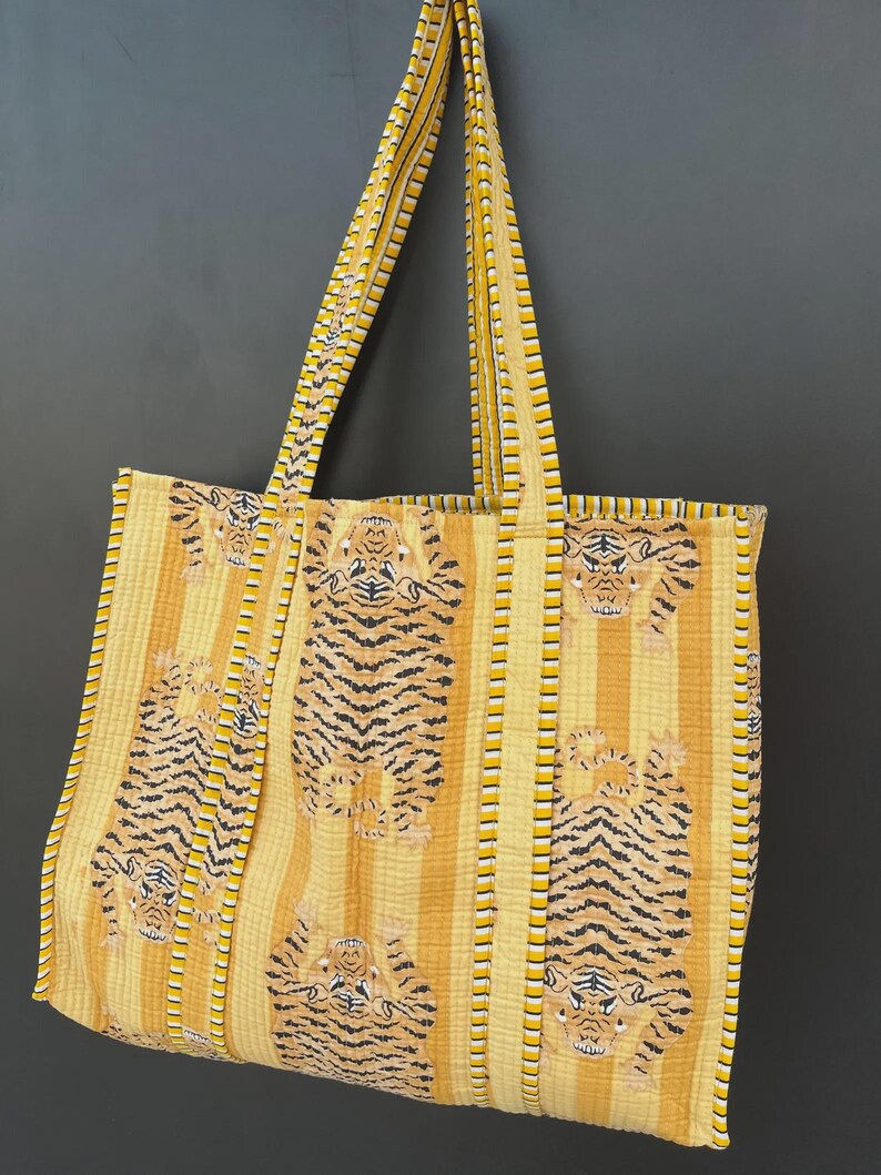 May include: A large tote bag with a yellow and orange striped pattern, featuring tiger illustrations. The bag has long straps and is made of a quilted fabric. The bag is designed for carrying items.