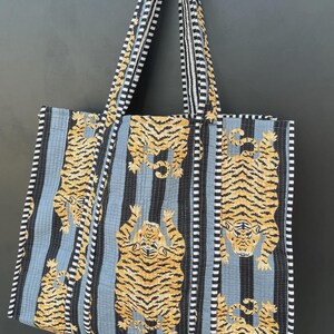 May include: A quilted tote bag with a repeating pattern of tigers in orange and black stripes against a light blue background. The bag has black and white striped handles and trim. The bag is made of fabric.